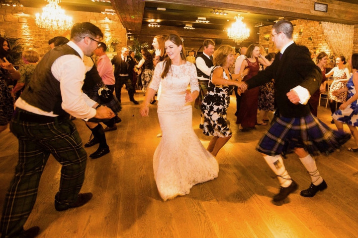 A bride and a guest in a kilt dancing a ceilidh with wedding guests at a Scottish wedding reception